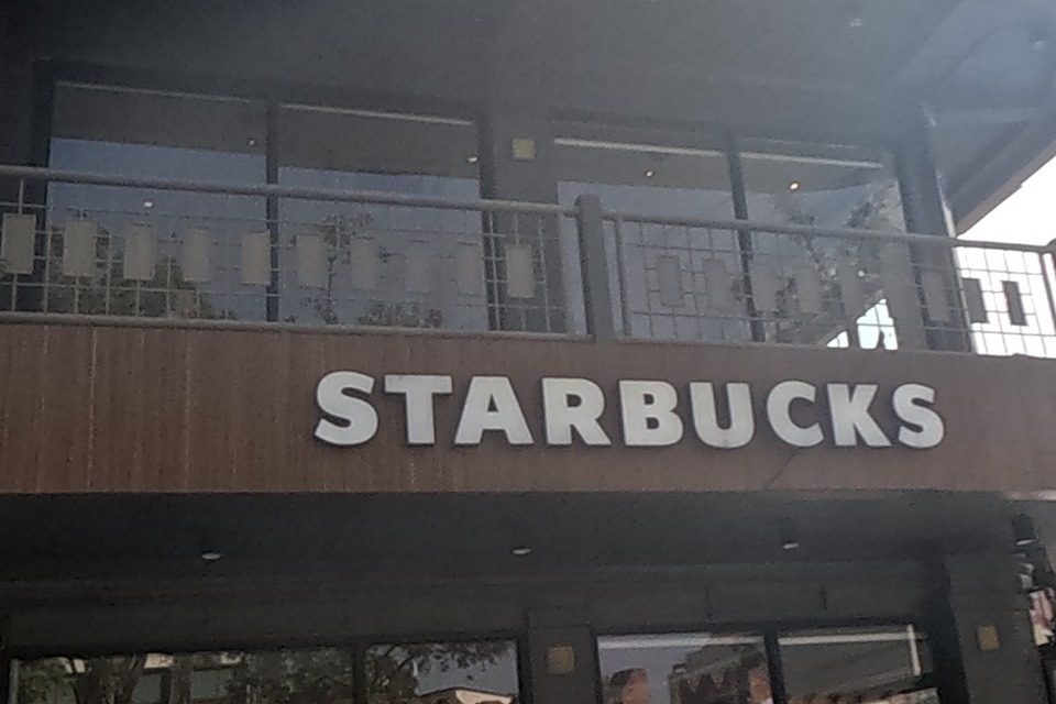A Starbucks with its green roundel logo occupies a modern wood-panelled corner building; 'K-perfect Nails & Spa Thaphae' is signed on the building next door; an old maroon pickup truck advertising day-trip tours (Tiger Kingdom sticker visible) and a scooter are parked outside, the red city wall reflected in the Starbucks windows.