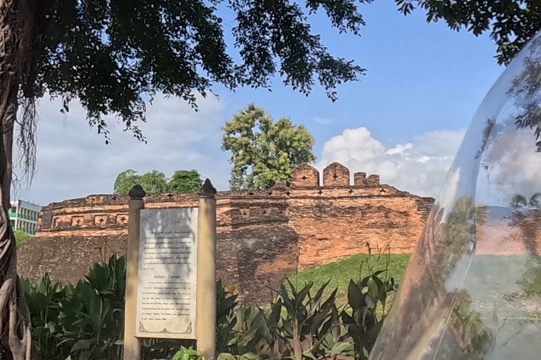 Through the trees, the red-brick ramparts of the old city wall's Chaeng Katam corner rise beyond the moat, with an old banyan tree and an information plaque on the near bank.