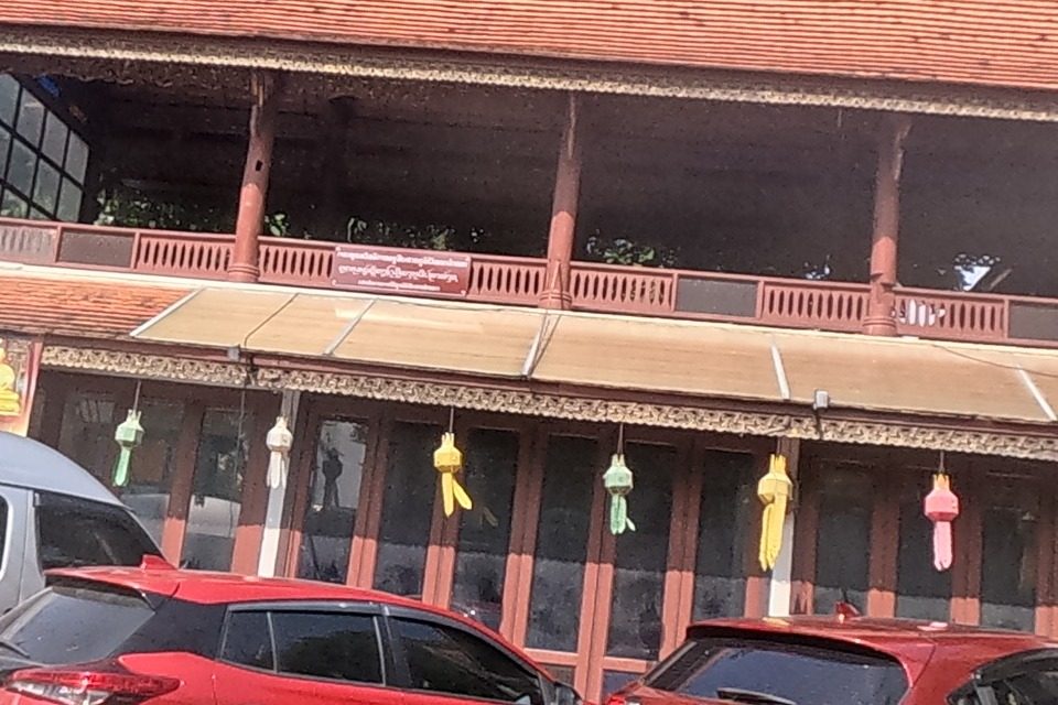 Wat Pansao's red-roofed viharn stands behind parked cars and a van, its carved gable and colourful paper lanterns hanging along the porch, a white chedi visible beside it.
