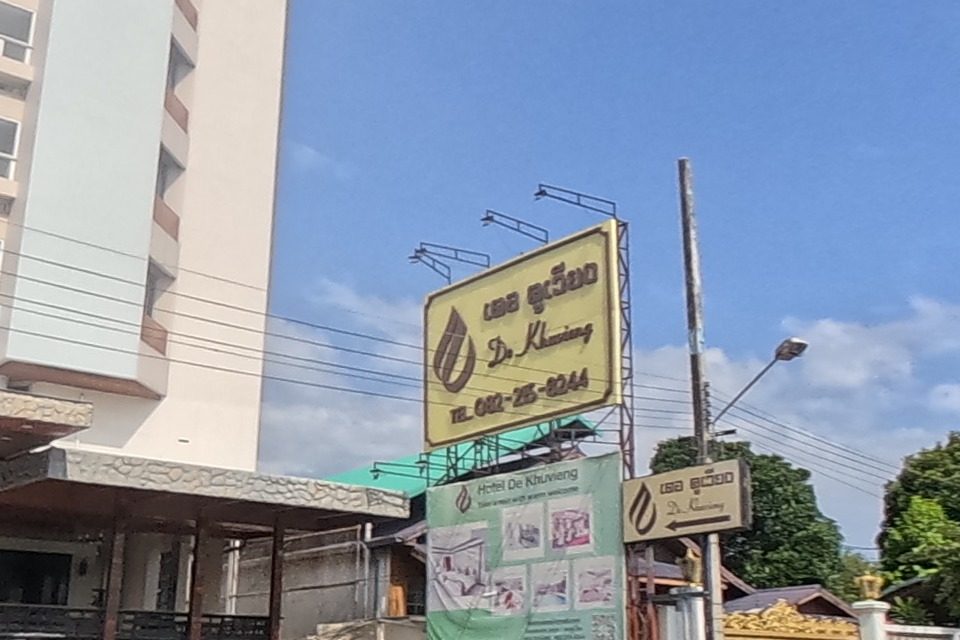 A yellow roadside sign clearly reads "De Khuvieng" hotel with phone number 082-215-8244, and a green banner below advertises "Hotel De Khuvieng" with room photos; further down the road a large purple building with a polka-dot facade and a big red/orange "P" logo is visible, which later frames confirm is a Power Buy electronics store.