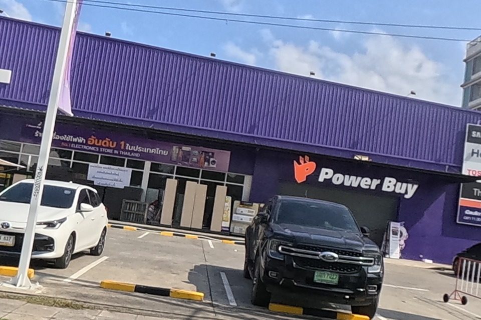 The Power Buy building's main entrance, its full name and "No.1 Electronics Store in Thailand" slogan legible along with Samsung, Haier, Toshiba and The 1 card logos; cars are parked in front.