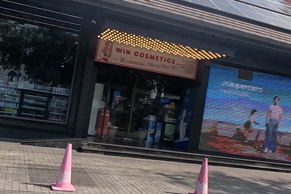 A modern black-fronted shop with a full solar-panel roof leads to “WIN COSMETICS” with a lit yellow marquee sign; pink traffic cones line the pavement.