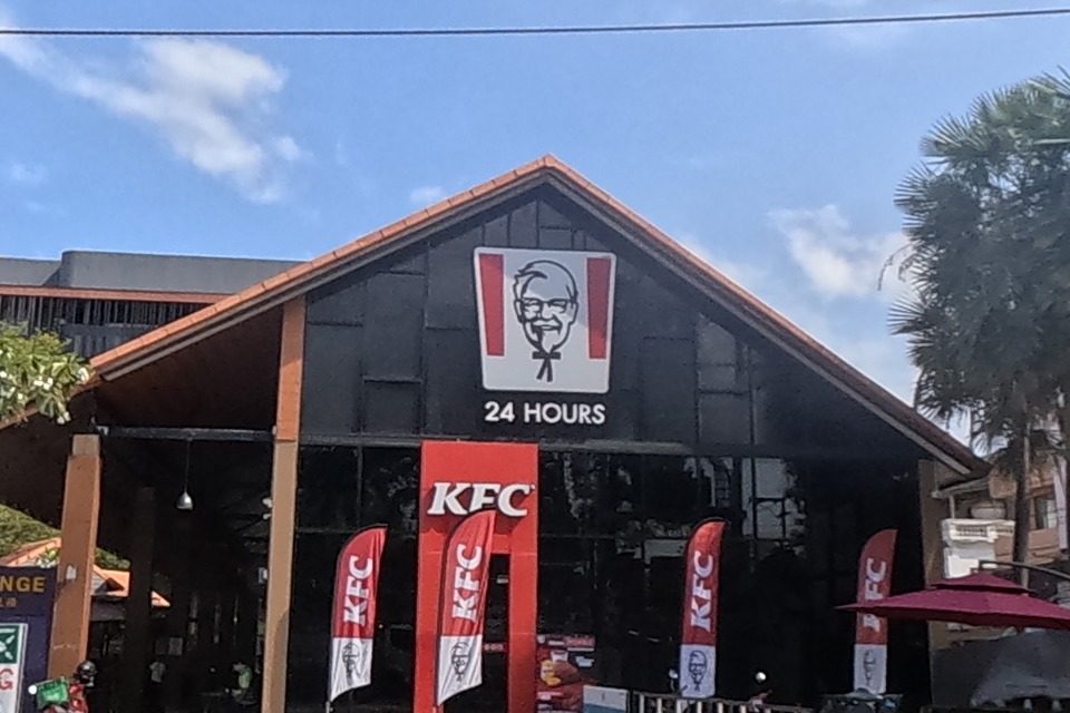 Pizza Hut (no. 1150) and KFC 24 Hours seen head-on, delivery riders' motorcycles parked between them; a banner advertises a Halal street food festival at Pun Hotel Chiang Mai.