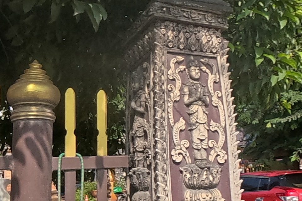 An ornately carved gate pillar with a relief of a standing devata figure marks the corner of the King Naresuan Shrine compound; a red car is parked on the sandy forecourt beyond the fence.