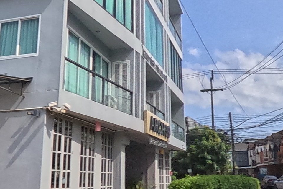 The grey “ARCH39” sign marks the corner of a modern building with teal-glass balconies; a blue motorcycle is parked on the patterned brick forecourt.