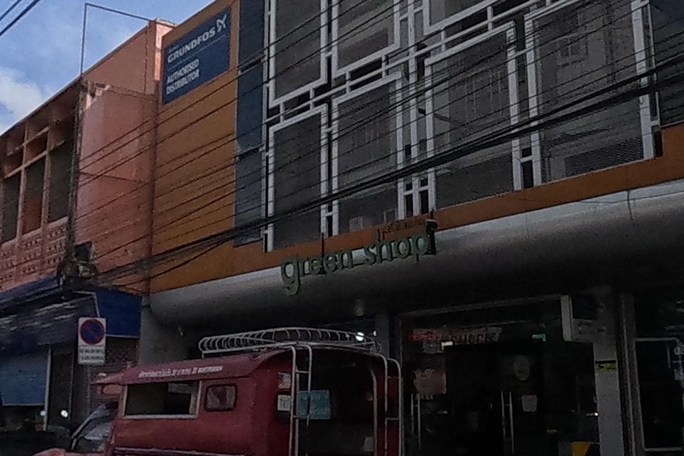 A shop called 'green shop' beneath an orange-panelled building advertises solar-panel installation to cut electricity bills, next to Grundfos pump-distributor signage and a parked red songthaew.