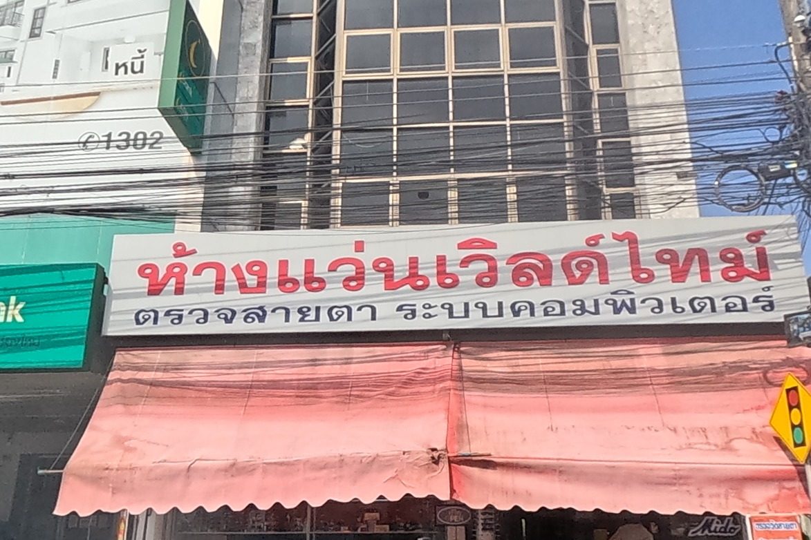 An eyewear shop with a large red sign advertises computerised eye exams next to an Islamic Bank of Thailand branch under a green fascia; motorbikes and a silver sedan are parked outside.