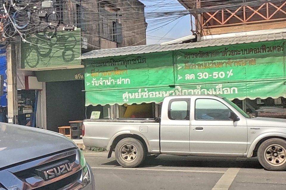 A green-signed watch repair centre advertises computer-tested repairs, pawning service and 30-50% discounts, beside a currency/gold exchange sign; a silver pickup and an SUV are on the road, and a delivery rider passes on a red motorbike.