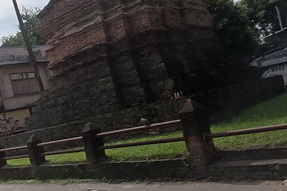 The brick ruin of an old chedi, believed to be Wat Pan Sat, stands behind a low brick-and-iron-rail fence on a grass lawn, opposite parked cars.