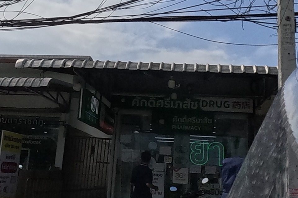 A glasses shop 'แว่นตาราชภัฏ' and a pharmacy 'ศักดิ์ศรีเภสัช DRUG' with a lit 'ยา' sign sit side by side beneath a shared awning, a customer at the pharmacy counter.