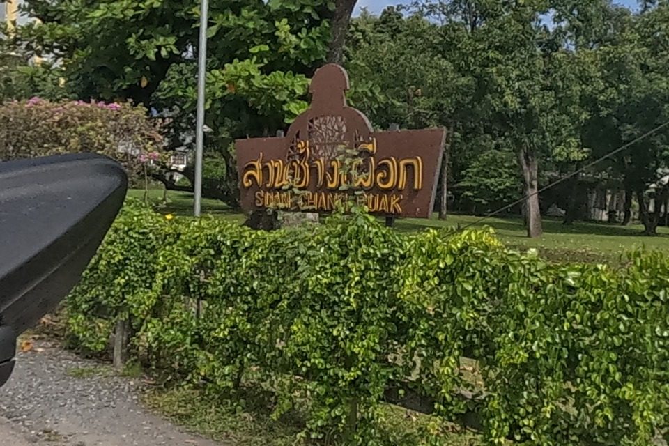 A wooden sign reading 'สวนช้างเผือก / SUAN CHANG PUAK' marks the entrance to a shaded public park behind a hedge, with a curved pavilion and parked cars visible down the drive.