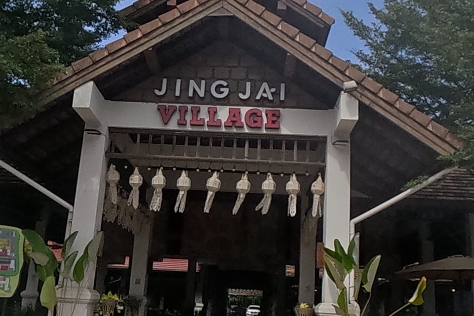The grand teak-roofed gateway of 'JING JAI VILLAGE' frames a red-carpeted entrance hall, flanked by 'ROGU ROTI' and other market shops, a green Grab discount stand out front.