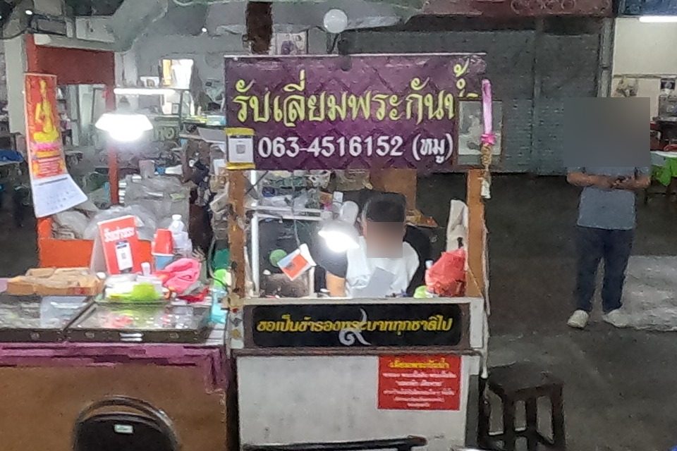 The หมู casing stall with its purple sign and phone, a man working at the lamp; beyond, a red 'Amulet frame' sign with Chinese and a phone number.