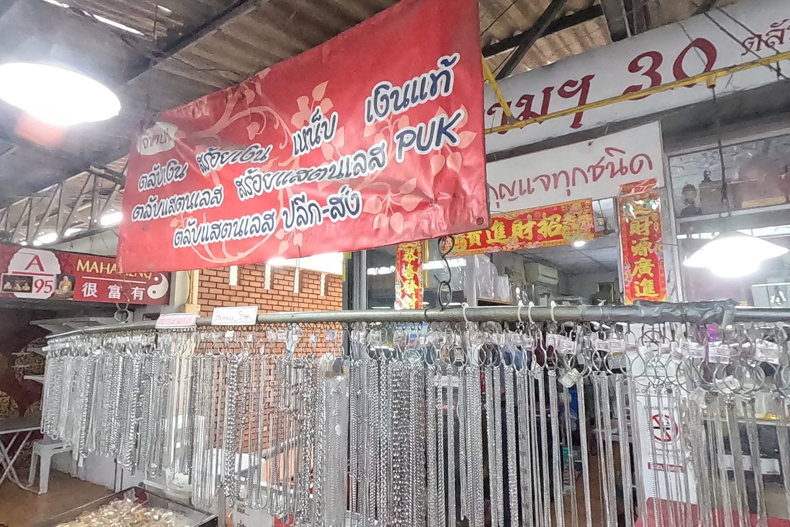 The PUK banner and the จตุคามฯ 30 sign above racks of chains and trays of cases, Mahaheng's red sign at the left.