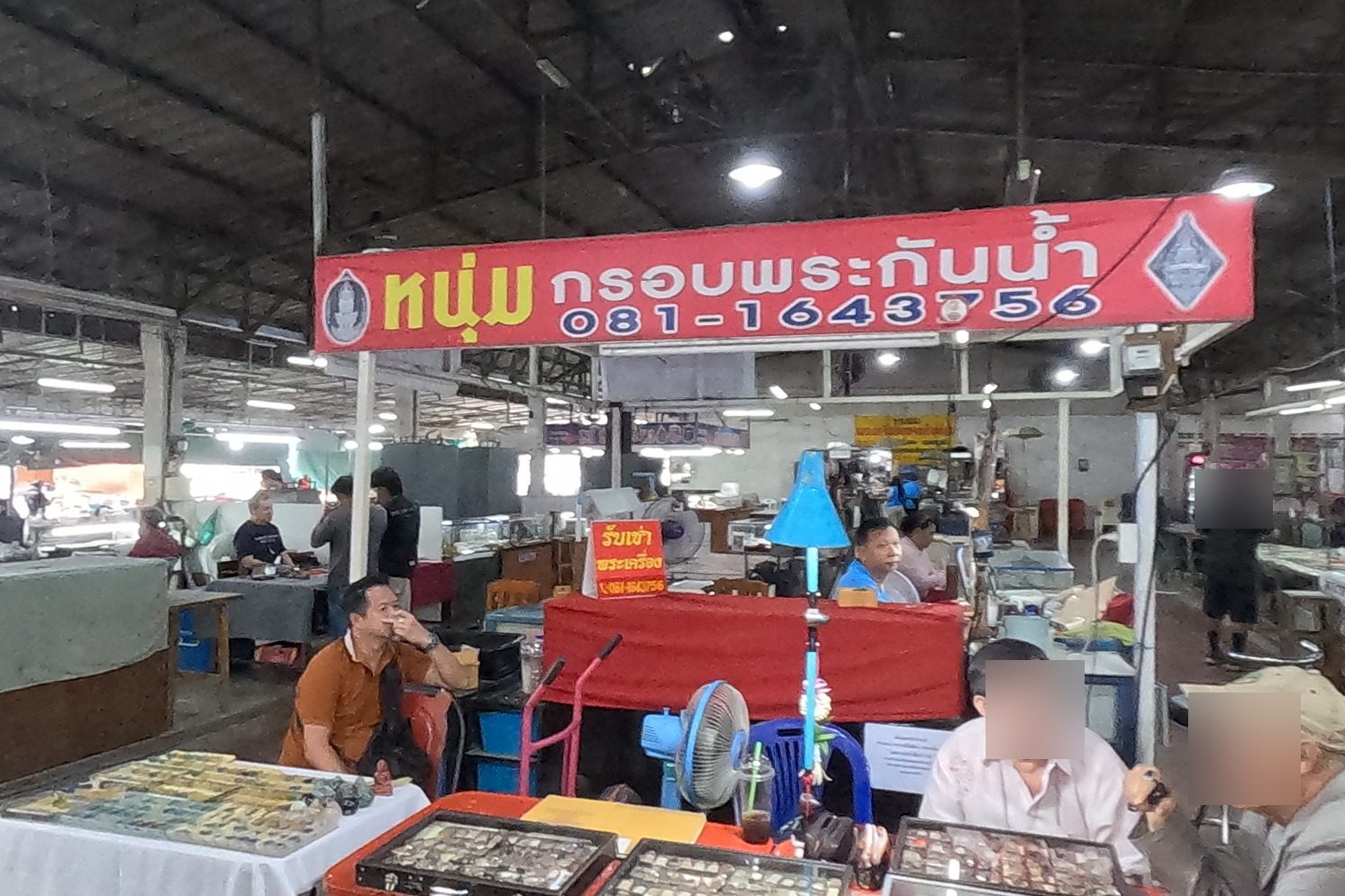 The หนุ่ม กรอบพระกันน้ำ stall under its red sign: dealers seated at a red table of glass trays of amulets, a white-cloth table and a blue lamp.