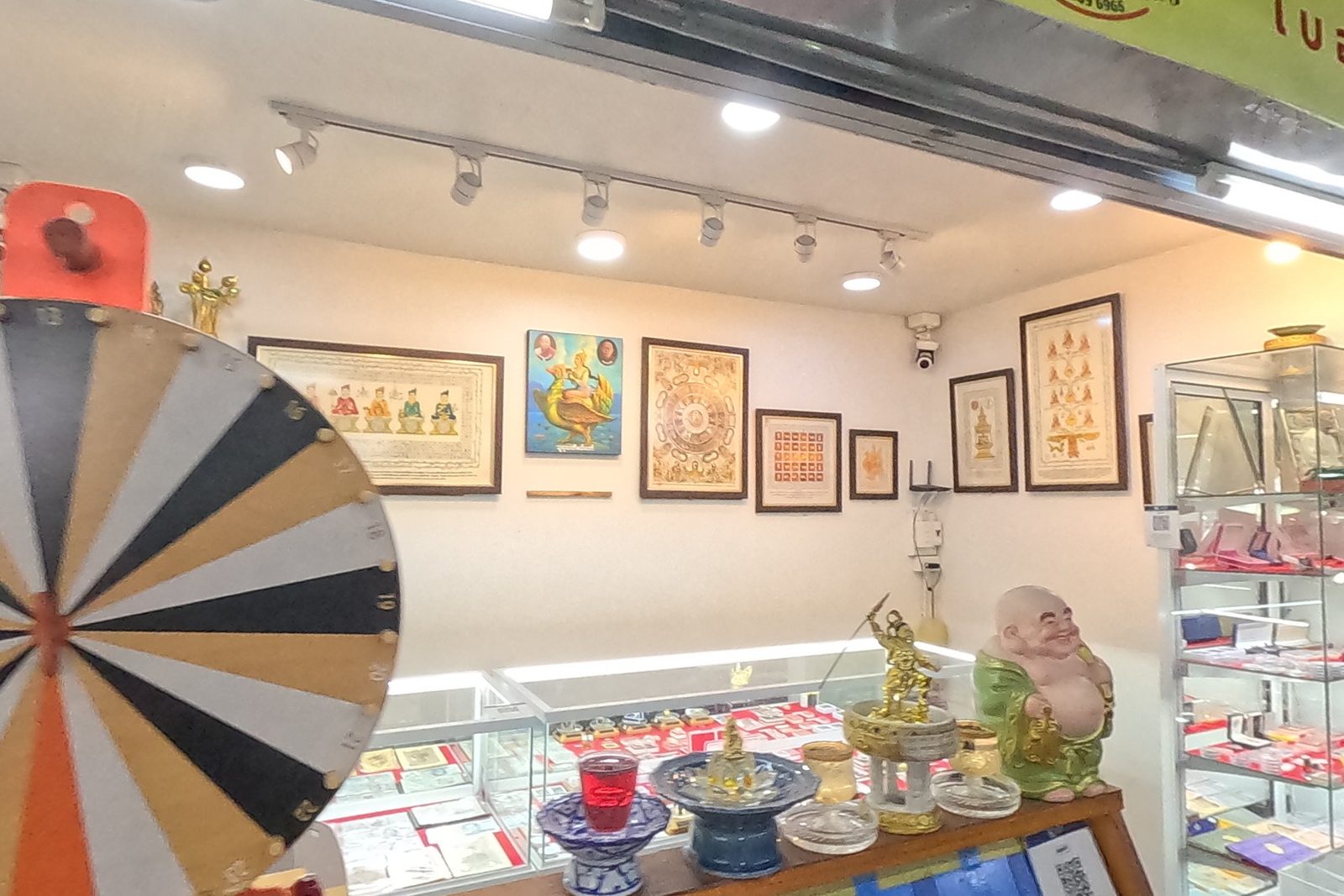 Inside TK-AMULET 65: a painted prize wheel, framed pictures, glass counters and a board of lottery tickets under the yellow-green fascia.