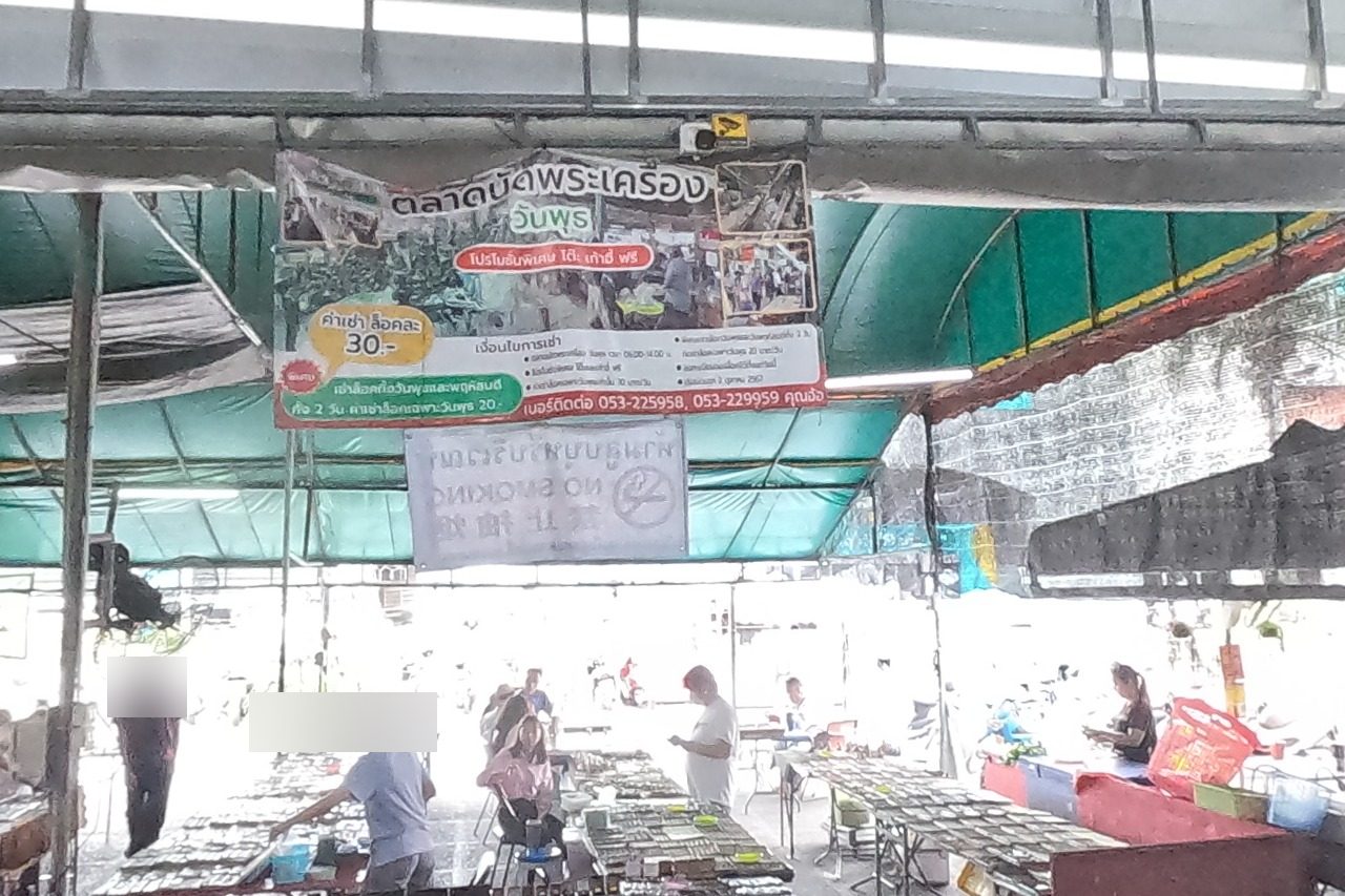 The open side of the hall under a green canopy: the amulet-market banner, rows of amulet tables in daylight, the khao soi stall at right and brass bowls on a curio case in front; a dealer in grey sits at his table of cased amulets.