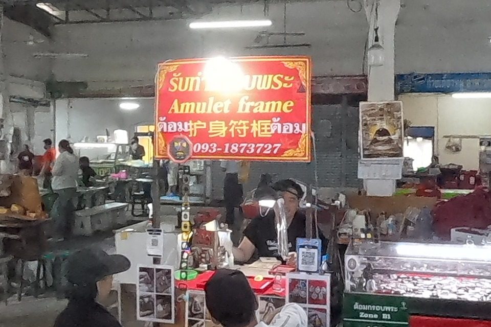A dealer inspects an amulet with a loupe beside bronze statues; the 'Amulet frame' stall of ต้อม glows behind.