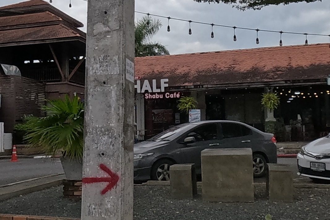 Lanna-style buildings with tiled roofs and string lights house HALF Shabu Café, with cars parked in the gravel forecourt and a red arrow painted on a concrete pole.