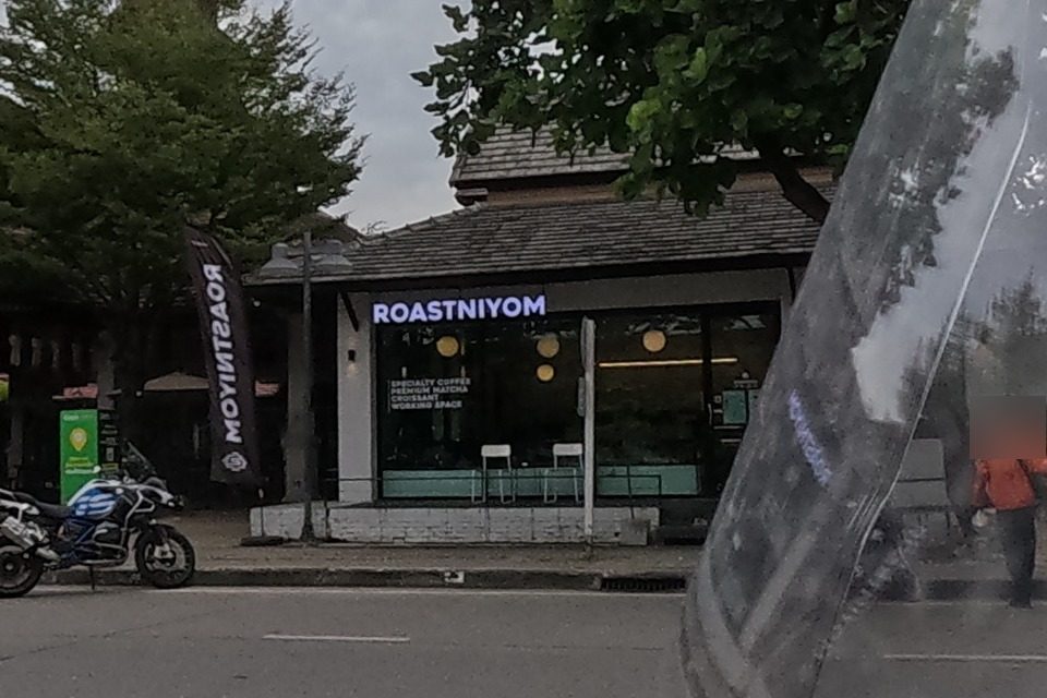 The Jing Jai Village gate and a specialty coffee and matcha shop called Roastniyom, with a person in an orange vest and parked motorbikes.