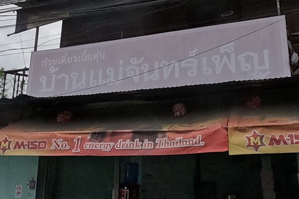 A stewed-beef noodle restaurant with tables set out front, under an M-150 energy-drink branded awning, motorbikes parked in front.