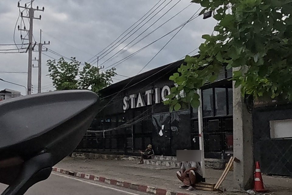 The dark 'STATION' building with people sitting outside on the kerb, next to a shophouse with a yellow awning and motorbikes parked along the road.