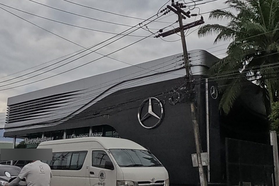 A black Mercedes-Benz dealership building with the Charoen Motor sign, a palm tree beside it, cars and a motorbike on the road.