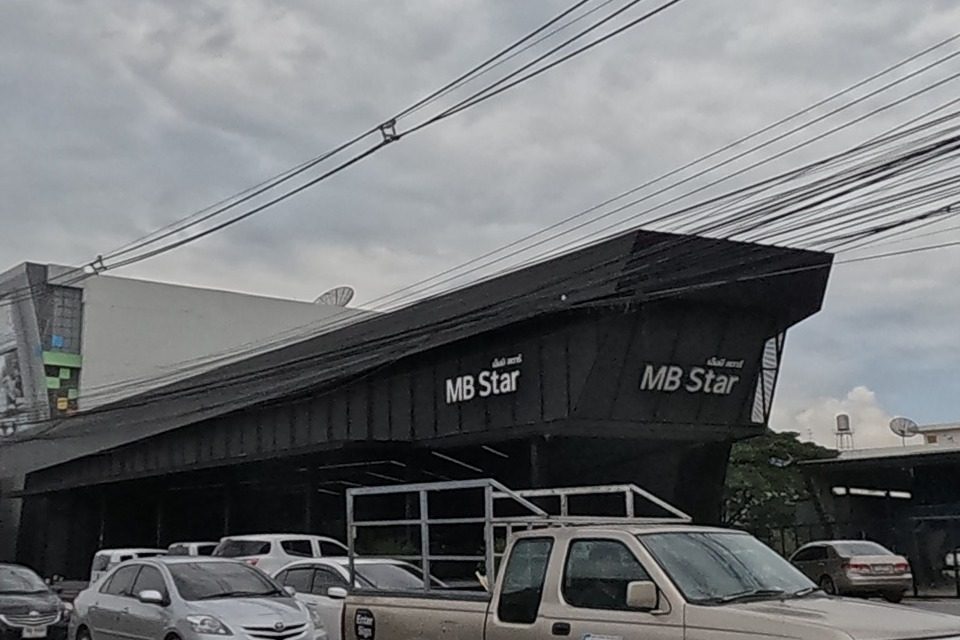 A black warehouse-style building with an 'MB Star' sign (Mercedes-Benz parts/service), a beige pickup with a Lalamove delivery sticker and other cars on the road.