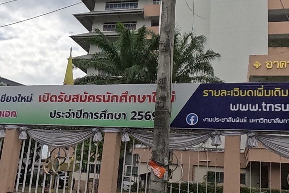 The fenced entrance of Thailand National Sports University, Chiang Mai Campus, with Thai flags, a royal emblem, and a banner announcing new-student admissions for bachelor's, master's and doctoral programmes (academic year 2569), website www.tnsucmi.ac.th.