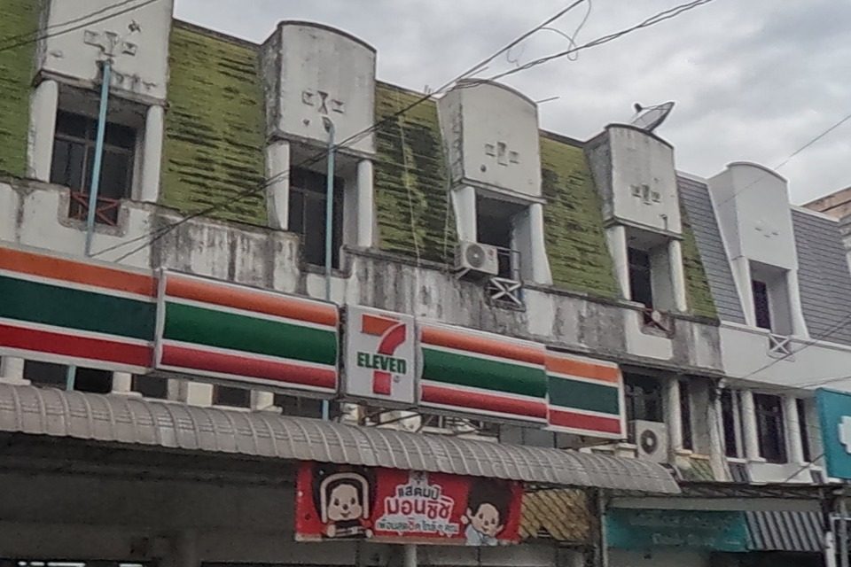 A 7-Eleven store with its red-green-white sign sits at a corner, next to 'Daily Dose Pharmacy' and a yellow children's-wear shop sign; a barbershop sign for '120 baht haircuts' is visible further along.