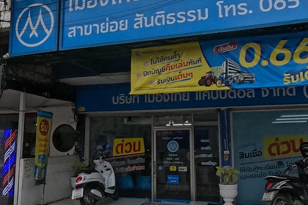 A two-storey shop signed 'เมืองไทย แคปปิตอล จำกัด (มหาชน) สาขาย่อยสันติธรรม' (Muangthai Capital PCL, Santitham sub-branch) advertises fast loans at 0.66% with car-registration and land-title collateral, phone 065 525 7566; a barbershop 'ตัดผมชาย 120' sign stands beside it.