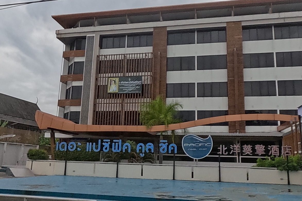 The Pacific Cool Chic hotel, signed in Thai, English ('เดอะ แปซิฟิค คูล ชิค / PACIFIC COOL CHIC') and Chinese ('北洋葵摯酒店'), rises behind an empty blue-floored plaza with a round hotel sign and a sand-for-sale notice at the roadside.