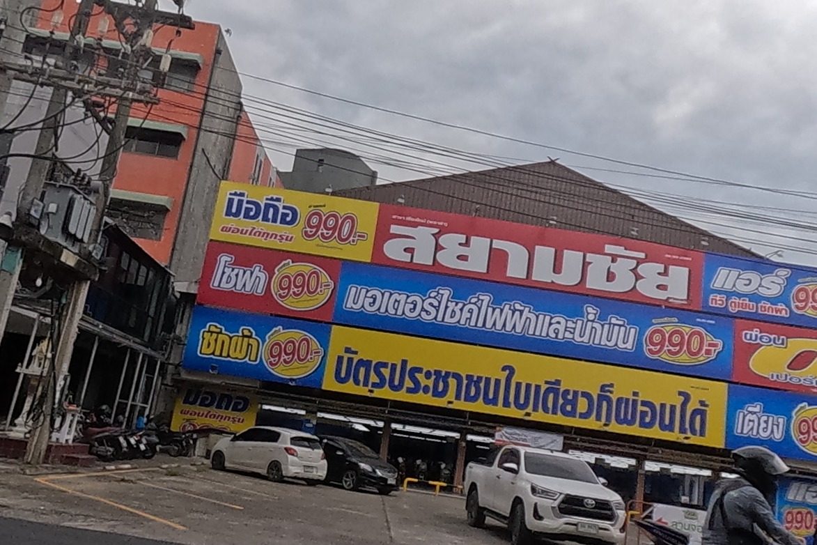 A large installment-retailer billboard for 'สยามชัย' advertises mobile phones, air conditioners, sofas, motorcycles, washing machines and beds all at 990 baht instalments, approved with just an ID card, over a car park with several vehicles; an 'ICON PARK' sign is visible on a building beyond.