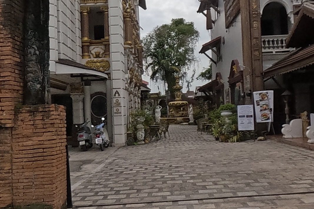 An ornate brick gateway with gold trim opens onto a paved courtyard lined with opulent white-and-gold buildings, statues, potted plants and menu boards, likely the entrance to Versailles de Flore or the Pingdoi Hualin Boutique Hotel compound.