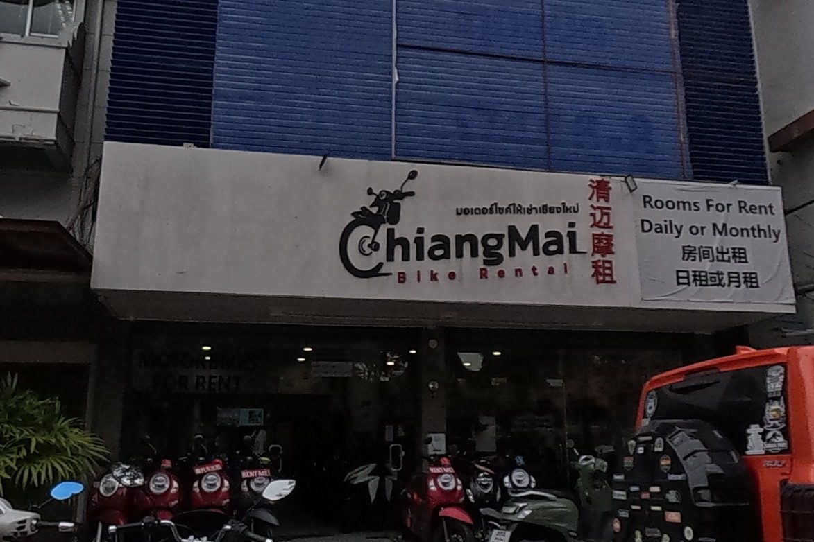 'ChiangMai Bike Rental' (清迈摩租), a motorbike-rental shop that also offers daily or monthly room rentals, displays rows of scooters and a Vespa parked outside; a bird's-nest health-product shop sign is visible above on the corner.
