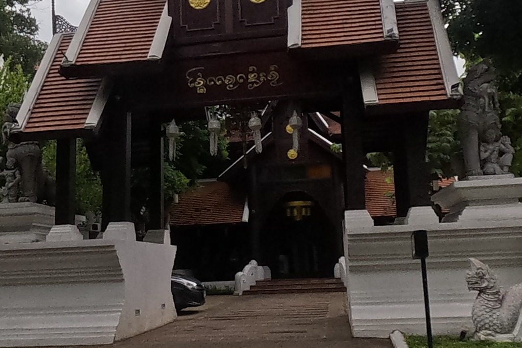 An elaborate Lanna-style tiered gateway, flanked by white stucco singha guardian statues and small white chedis, marks the entrance to 'The Rim' resort, its stone sign reading 'RIM' at the roadside.