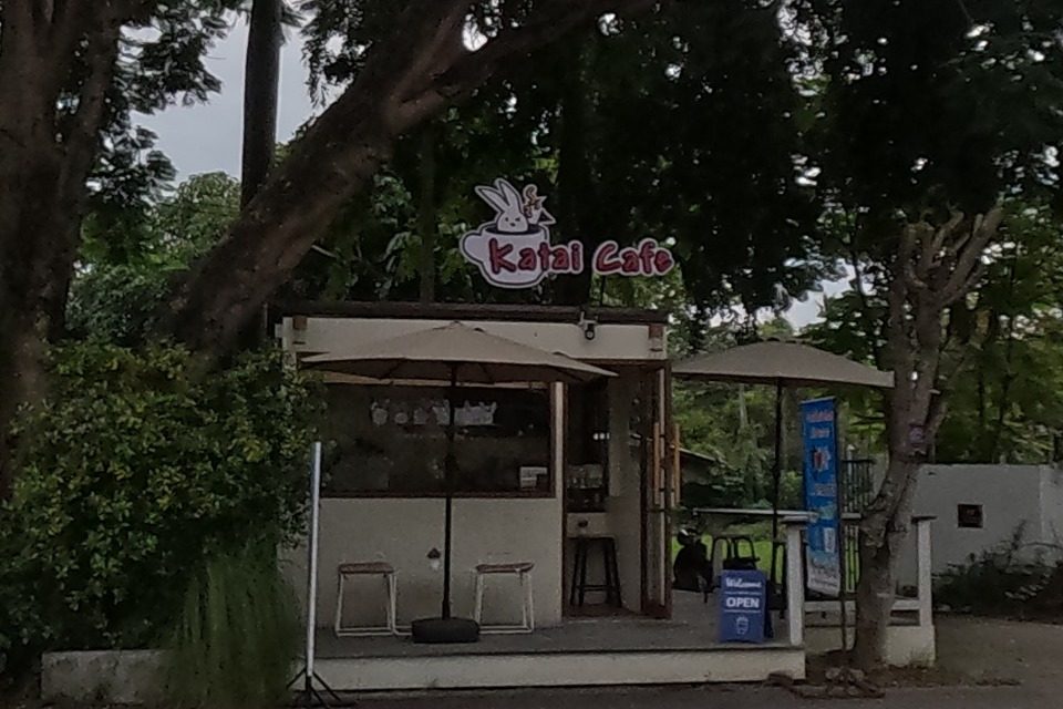 A small free-standing kiosk cafe, Katai Cafe, with outdoor seating under umbrellas beside a noodle shop sign advertising Coca-Cola.