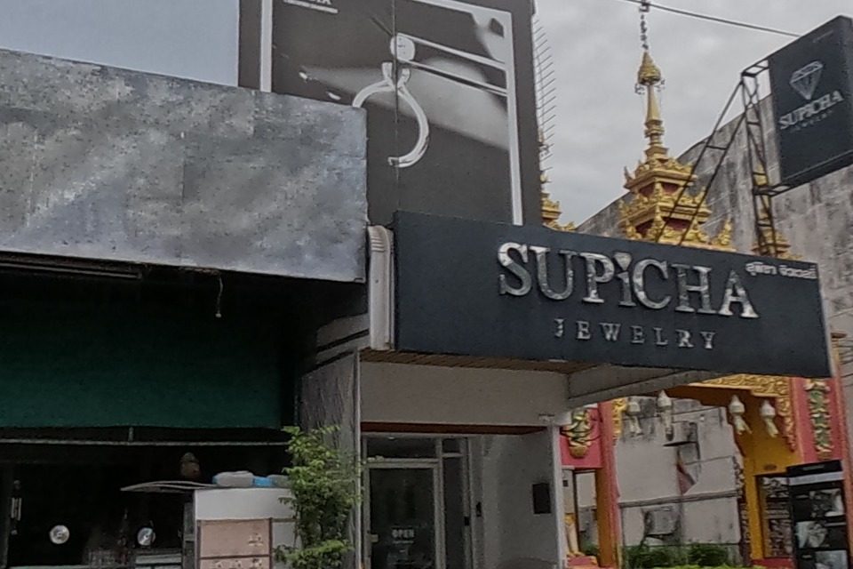 A gold-buying shop (หลอมทอง ล้านนา) advertising cash for gold, rose gold and silver, next to Supicha Jewelry with a GIA diamond ad; a red 'Ranger' branded pickup parked outside.