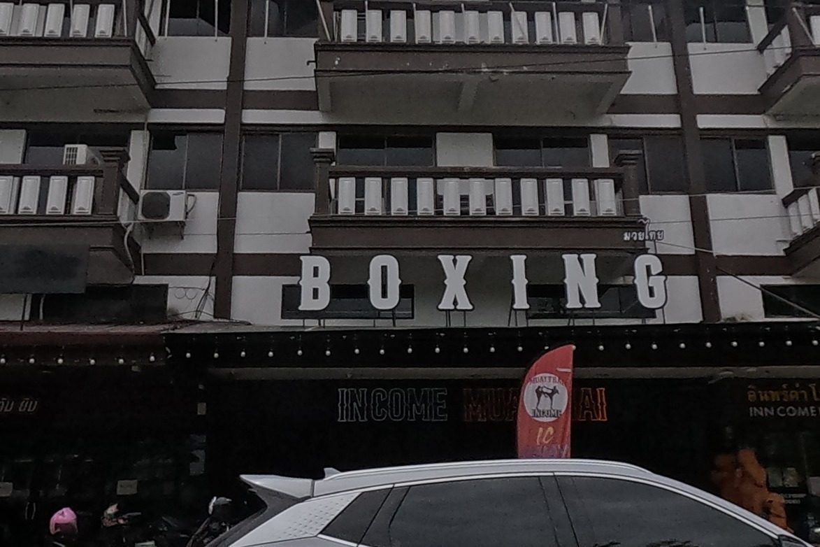 A muay thai gym with a large 'BOXING' sign, scooters and bicycles parked outside, a hostel next door.
