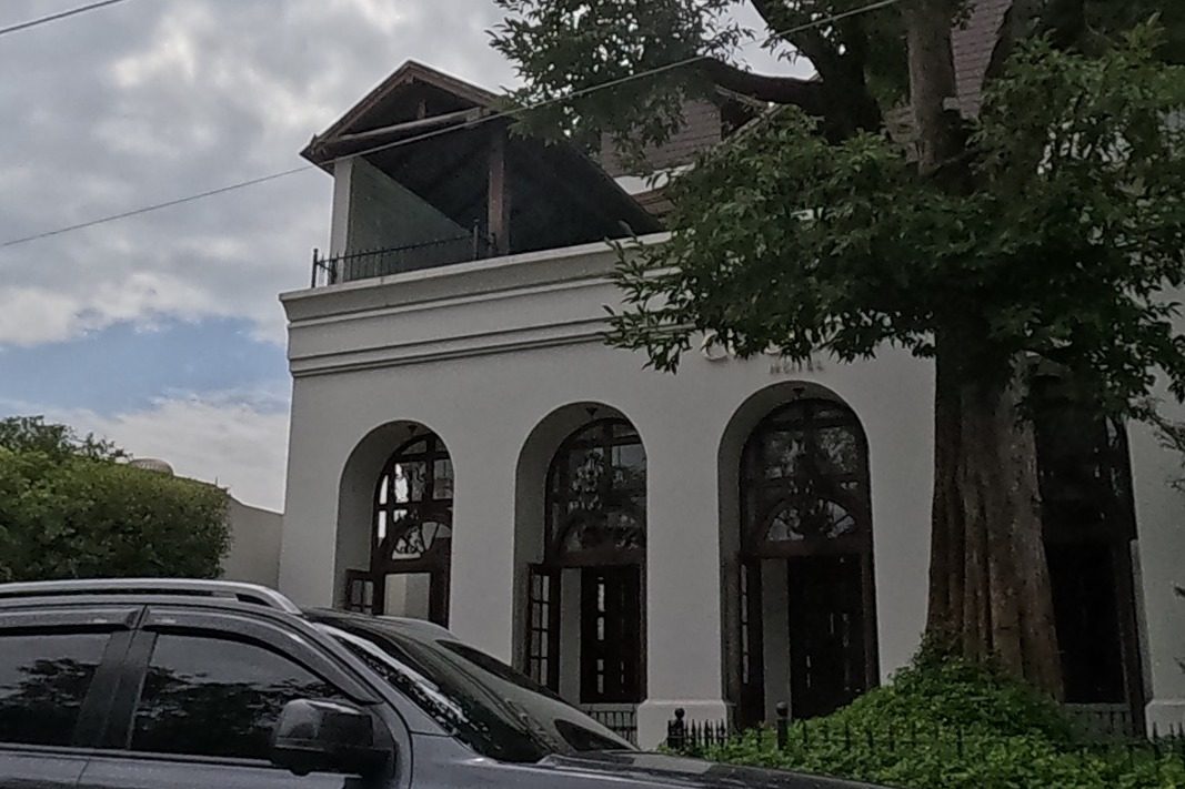 A white colonial-style hotel building with arched windows under a big tree, a beauty accessory shop next door.