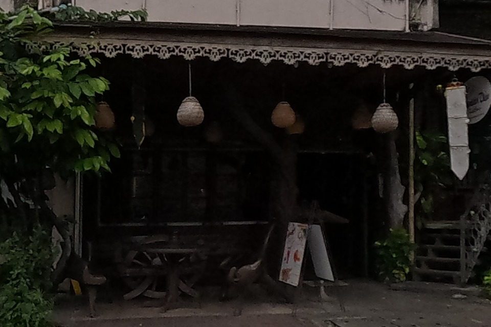 A shaded cafe with round paper lanterns under an ornately carved wooden eave, plants framing the entrance.