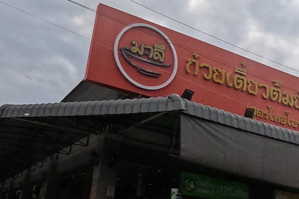 A blue Krungthai Bank ATM kiosk and a green ATM booth stand before a parking area leading to a noodle restaurant signed 'Malee'.