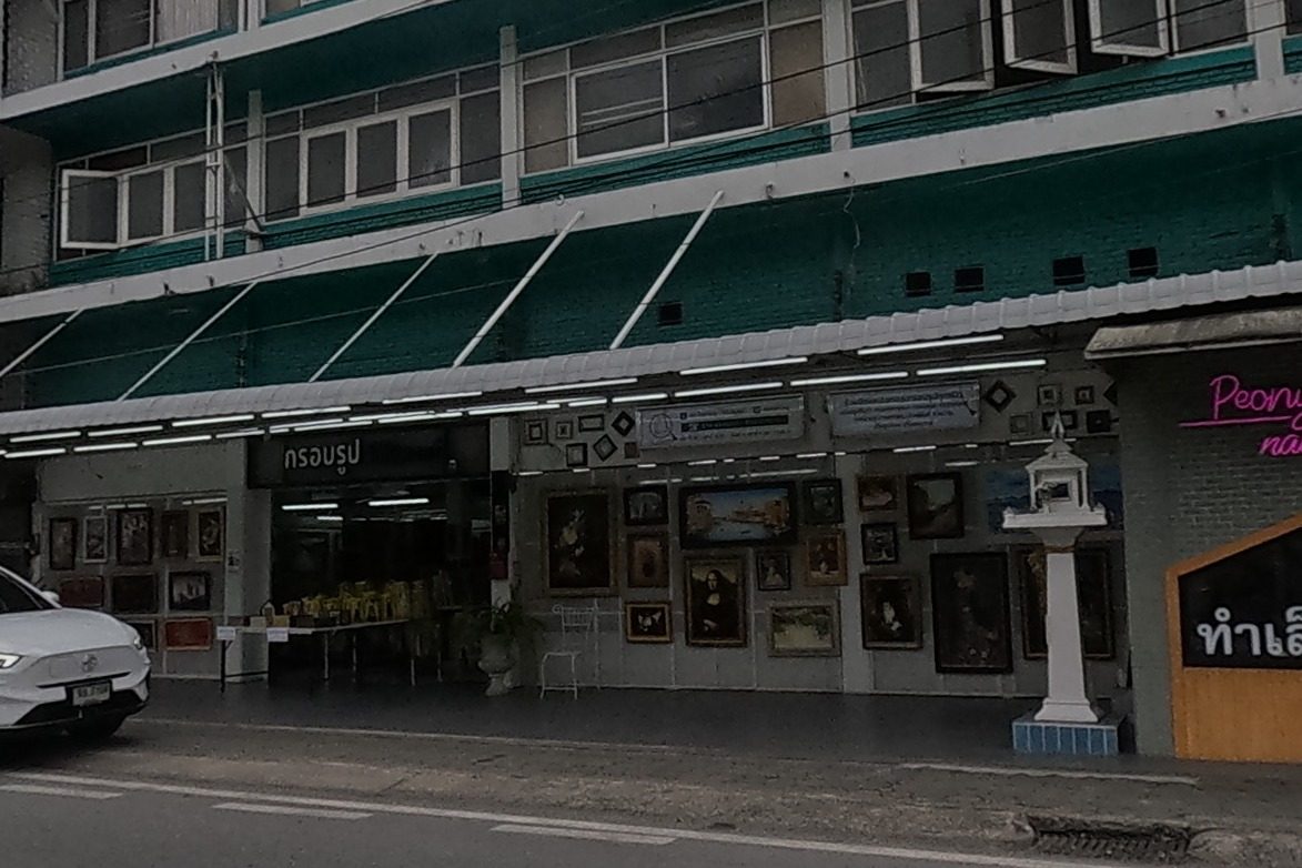 A picture-framing shop hung with portrait paintings sits beside a nail salon, with a small white spirit house on a pedestal out front and a scooter passing.