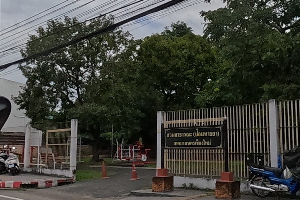 A public park with a playground behind a slatted fence has many scooters and motorbikes parked along the curb.