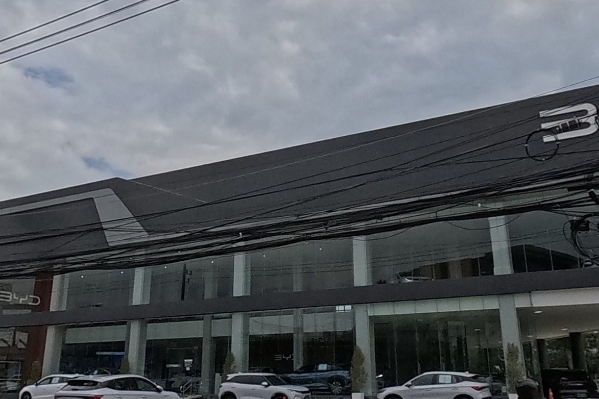 A dark modern BYD car dealership with a large glass showroom sits behind a black-and-white kerb, cars and scooters parked outside.