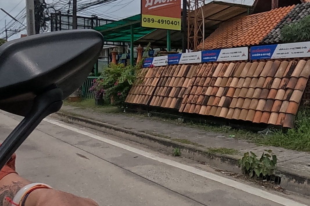 The rider passing a roof-tile yard with a red sign and a phone number strung overhead on wires.