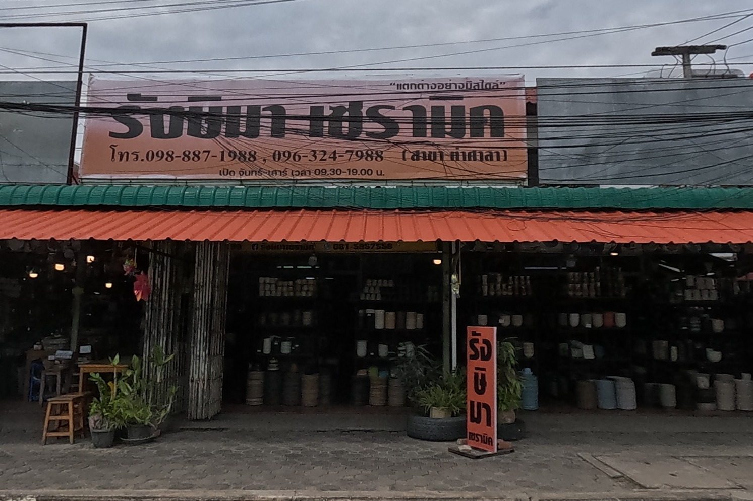 A large open-front pottery and ceramics shop with a banner reading its name, phone numbers and opening hours, a red sandwich board out front.