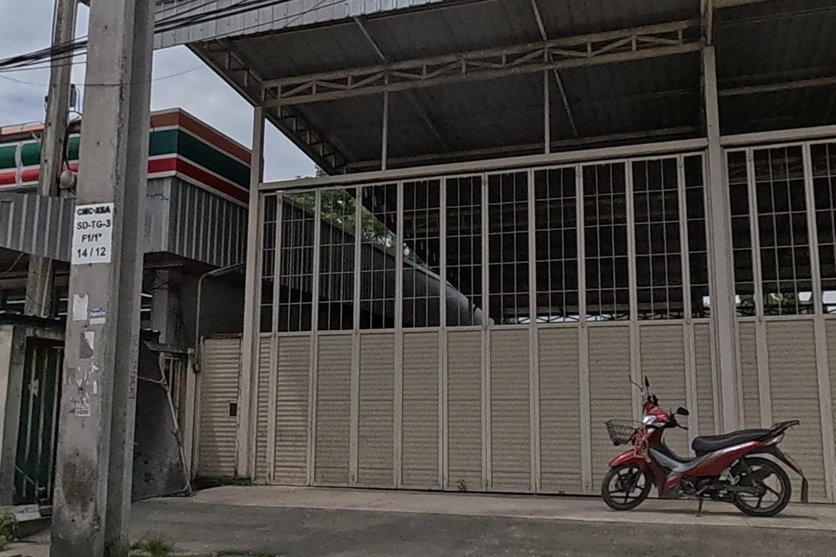 A 7-Eleven storefront next to a pharmacy sign reading 'ยา', a for-sale sign on a gated lot, and the noodle restaurant's red sign further down.