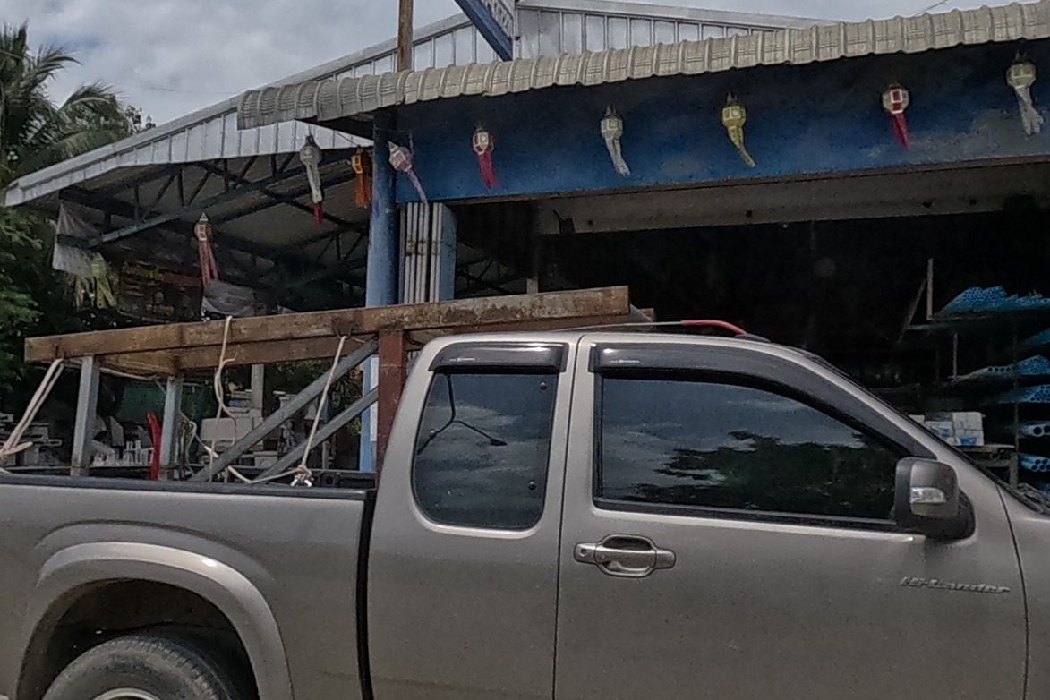 A pickup truck in front of a blue-fronted building-materials shop with keys and tools hanging over the open doorway, a small gold spirit house on a pedestal further along by a shed with a red pillar.