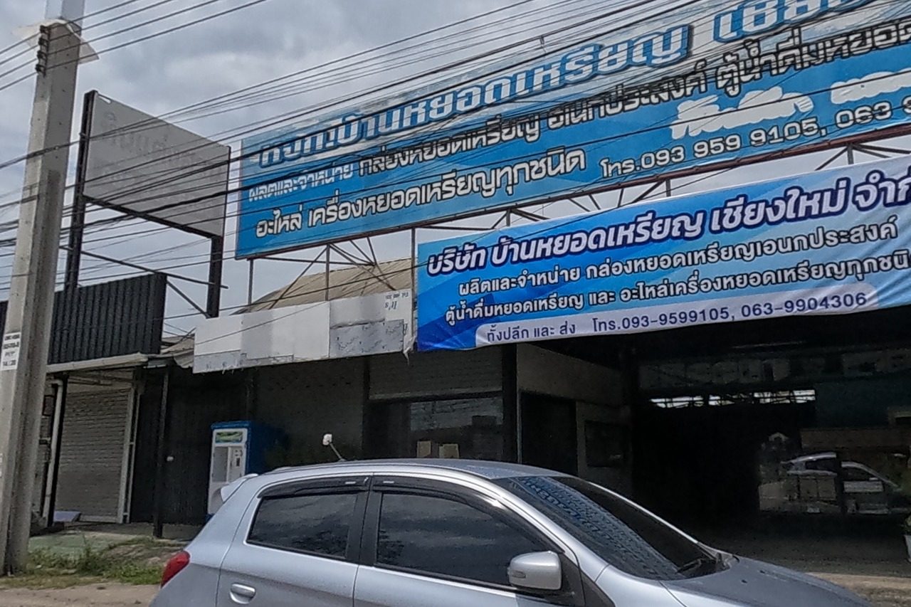 A coin-vending-machine company's banner-covered shopfront next to a fitness-equipment shop with a small shrine and a model house on stands out front, a scooter parked.